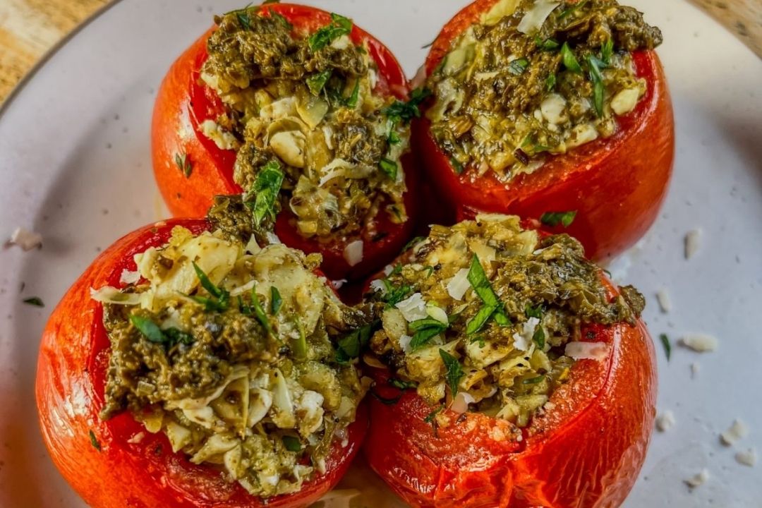 Four tomatoes on a plate, hollowed out and stuffed with a mixture of herbs, cheese, and possibly nuts, garnished with grated cheese and chopped herbs.