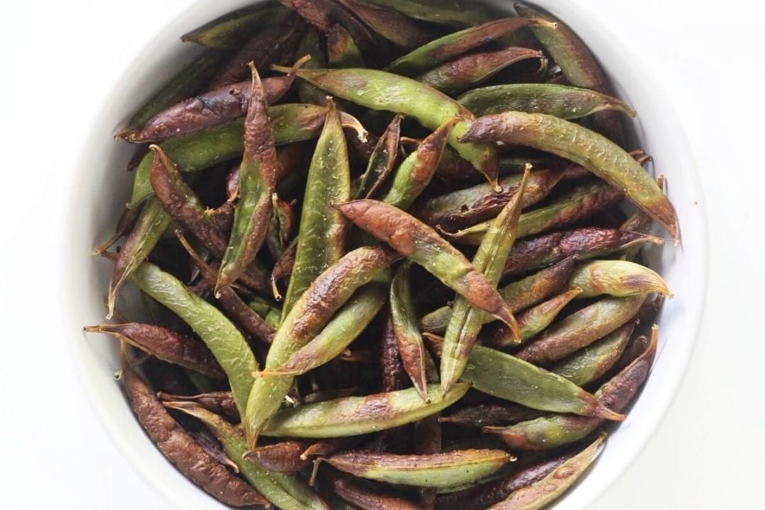 A white bowl filled with roasted green and browned snap peas on a white background.