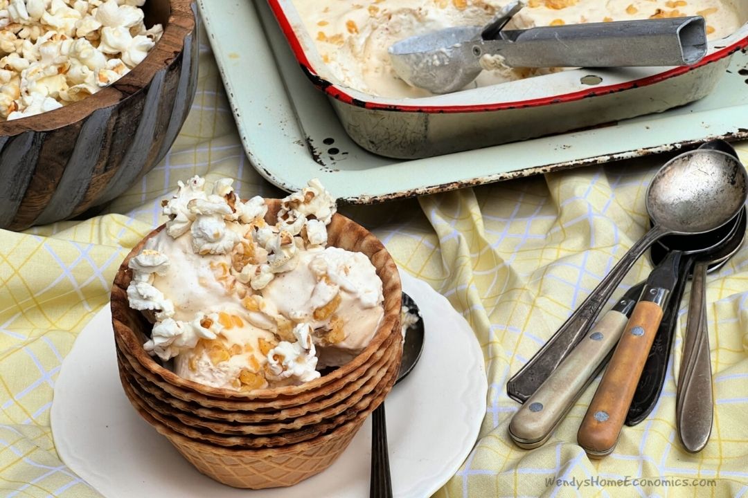 Waffle cone bowls with ice cream topped with popcorn and nuts, set on a plate beside an ice cream pan, scoop, and assorted spoons on a yellow patterned cloth.
