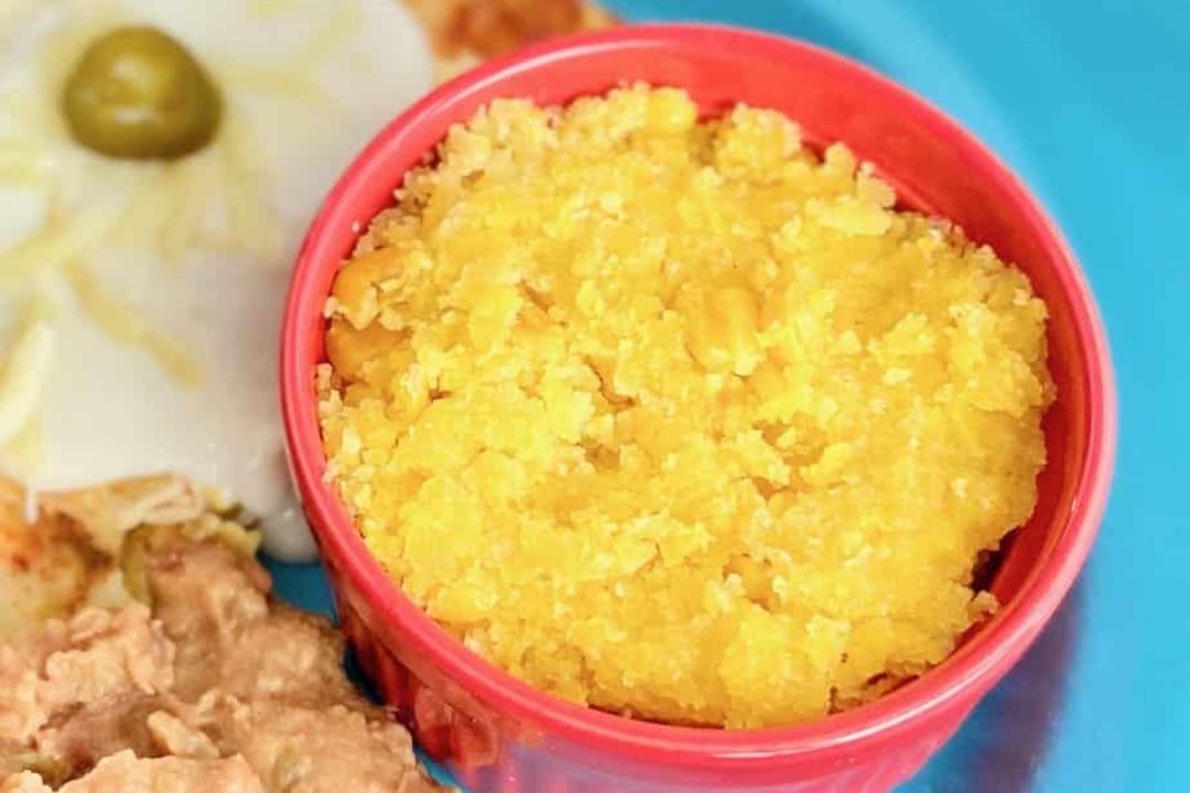 A red bowl filled with bright yellow corn pudding sits on a blue plate next to a fried egg, refried beans, and other food items.