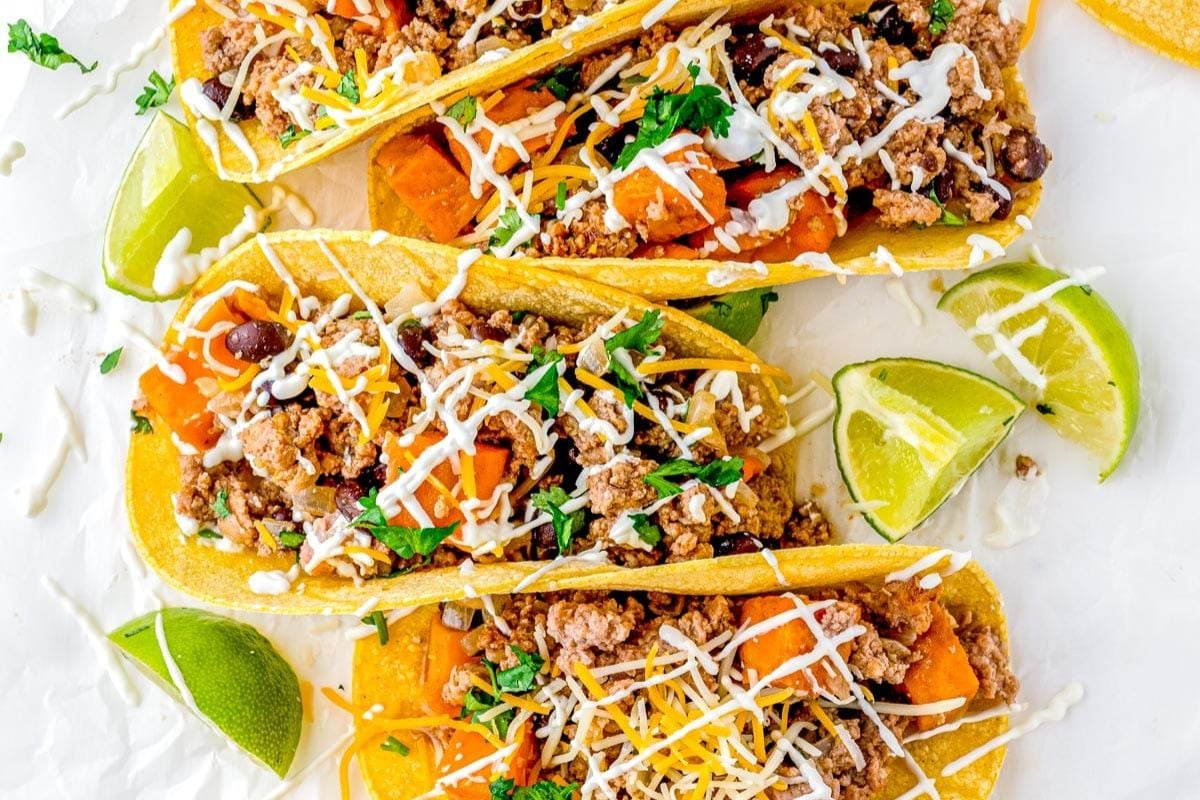 Three tacos filled with ground meat, beans, cheese, chopped herbs, and drizzled sauce, garnished with lime wedges on a white background.
