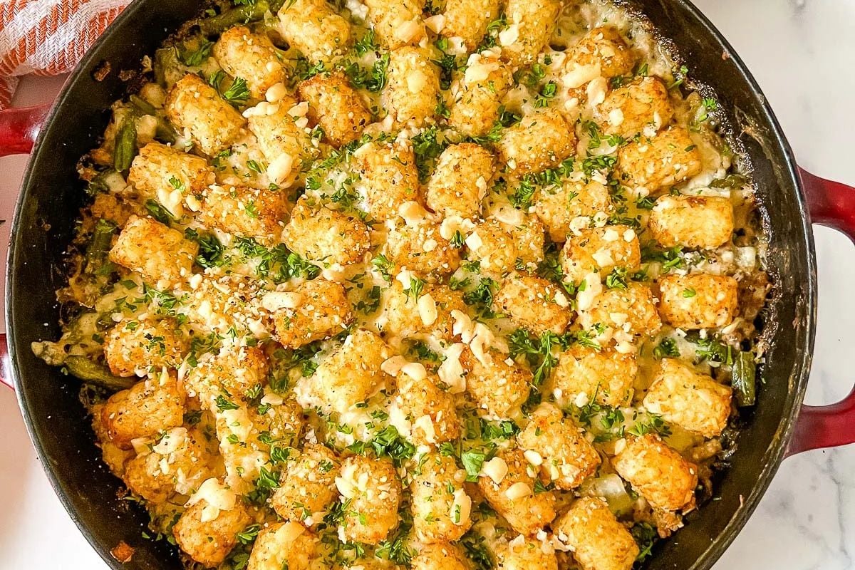 A skillet filled with tater tot casserole, topped with golden tater tots, chopped herbs, and crumbled cheese on a marble countertop.