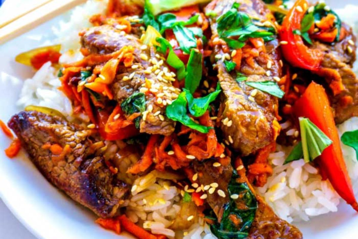 Beef stir-fry with sliced carrots, bell peppers, spinach, and sesame seeds served over white rice on a white plate.