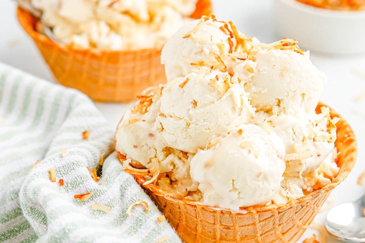 Scoops of vanilla ice cream with toasted coconut flakes served in a waffle bowl, next to a striped towel and a spoon.