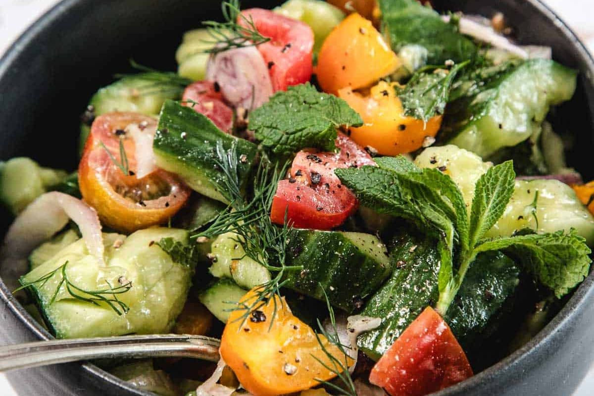 A bowl of cucumber and tomato salad with fresh herbs, red onions, and cracked black pepper.