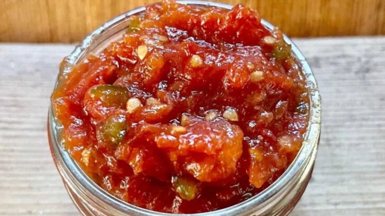 A close-up of a glass jar filled with chunky red tomato relish containing visible pieces of green pepper and seeds.