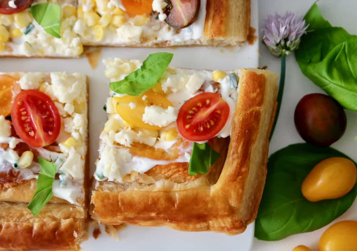 A close-up of a puff pastry tart topped with cream, cherry tomatoes, corn, feta cheese, and fresh basil, with basil leaves and tomatoes beside it on a white surface.