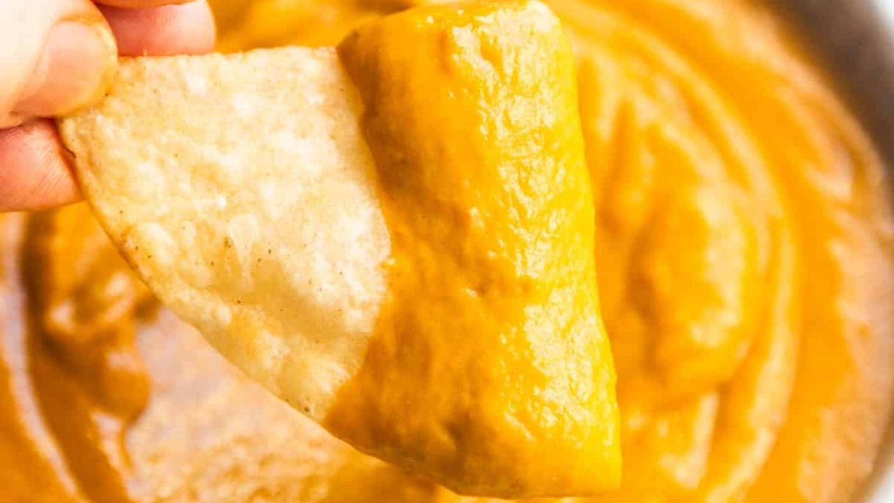 A hand holds a tortilla chip dipped in thick, orange cheese sauce.