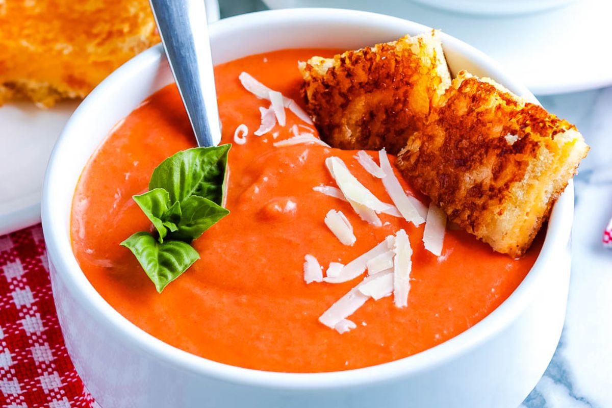 Bowl of creamy tomato soup topped with shredded cheese and basil, served with grilled cheese sandwich pieces, with a spoon in the bowl.