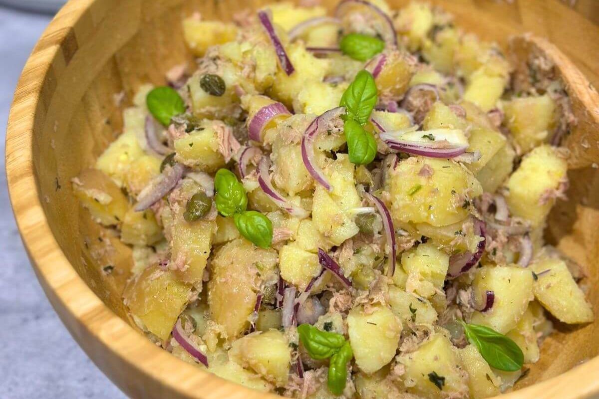 A wooden bowl filled with potato salad, red onion slices, capers, tuna flakes, and fresh basil leaves.