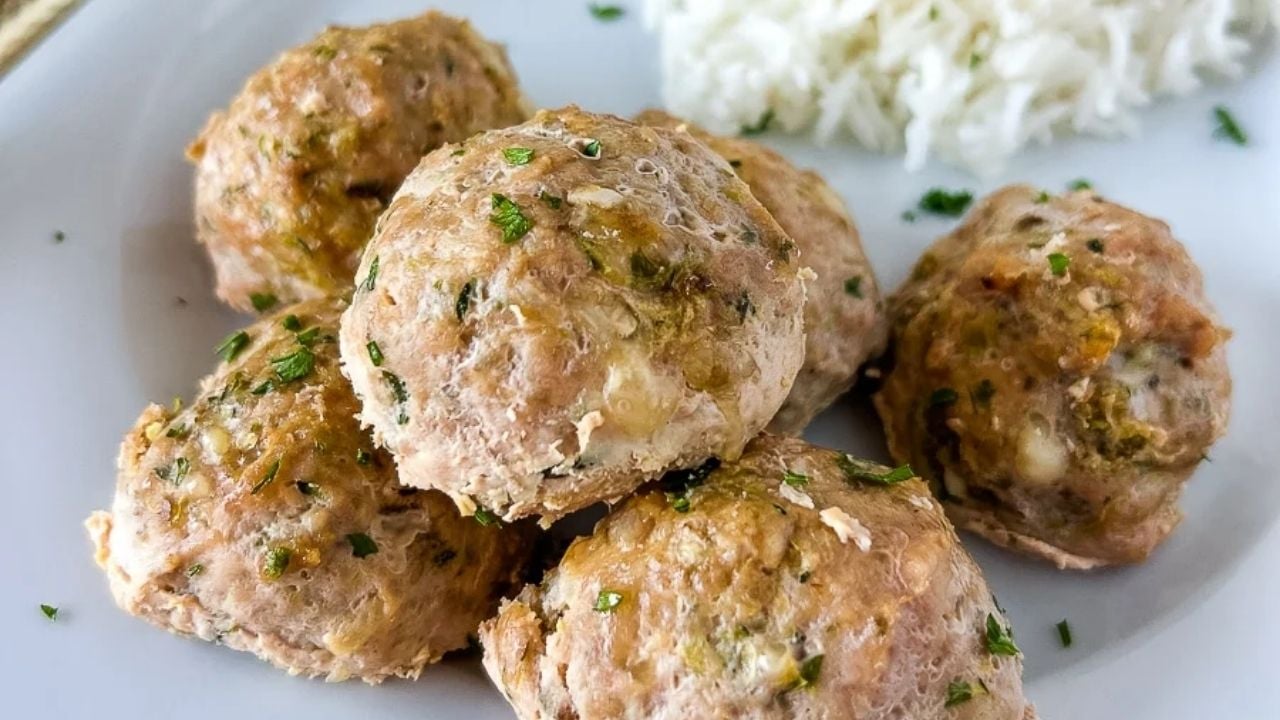 Five cooked meatballs garnished with herbs are served on a white plate, with a portion of white rice in the background.