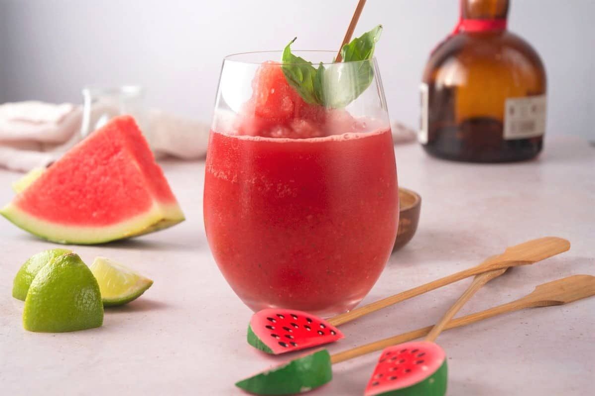 A glass of red frozen watermelon drink garnished with a basil leaf, with watermelon and lime wedges, cocktail sticks, and a liquor bottle in the background.