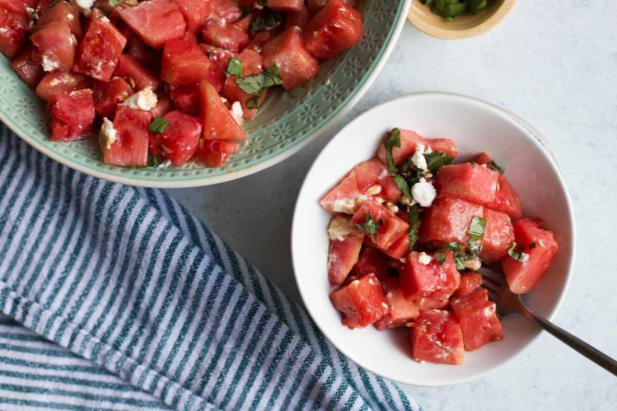 Two bowls of watermelon salad with feta cheese and herbs on a light surface, next to a blue-striped towel.