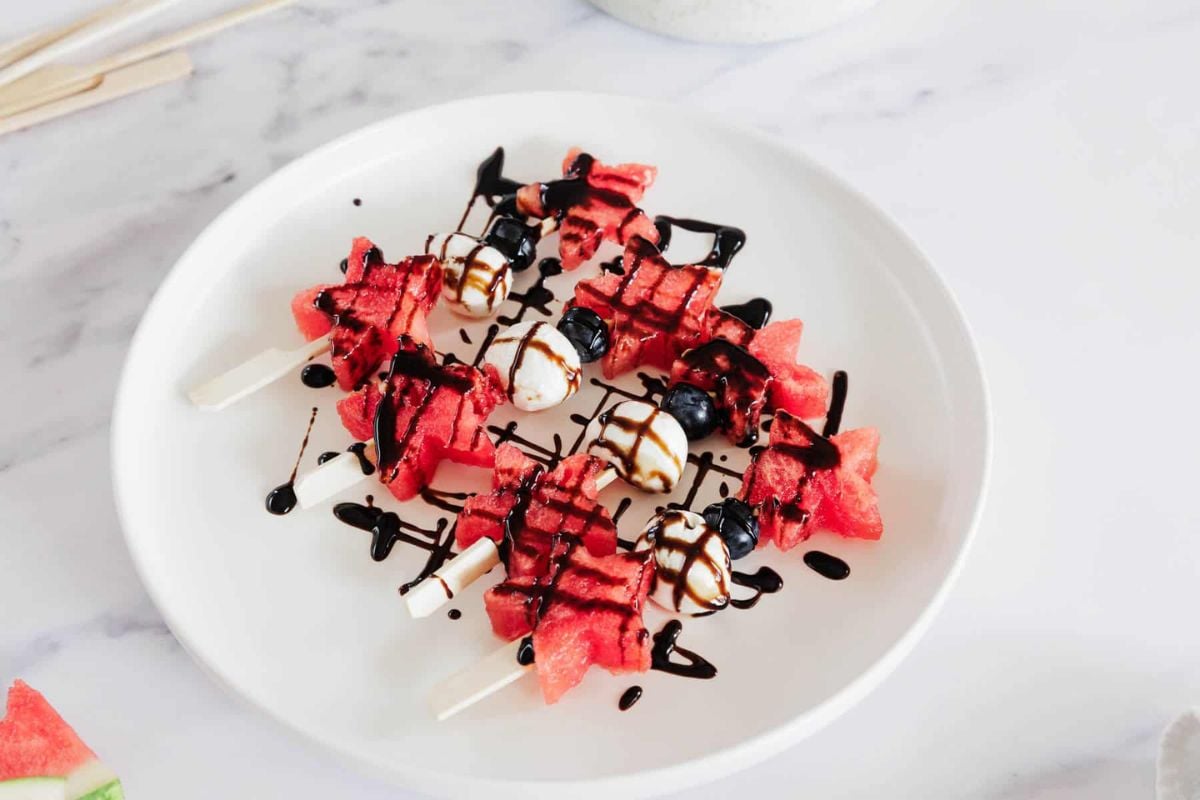 Watermelon and blueberry skewers topped with mozzarella balls and drizzled with balsamic glaze, served on a white plate.