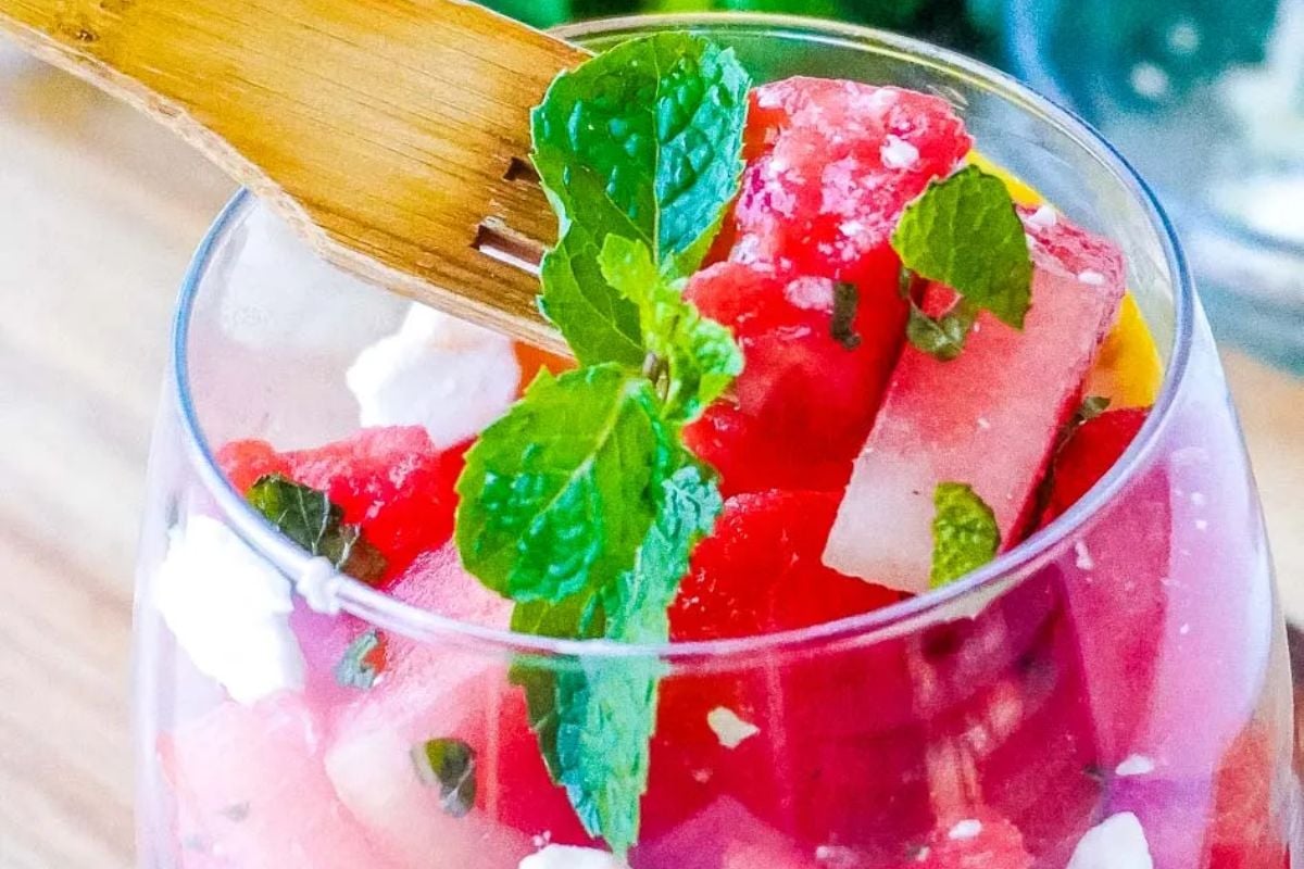 Close-up of a glass filled with watermelon cubes, feta cheese, and fresh mint leaves, with a wooden fork resting on top.