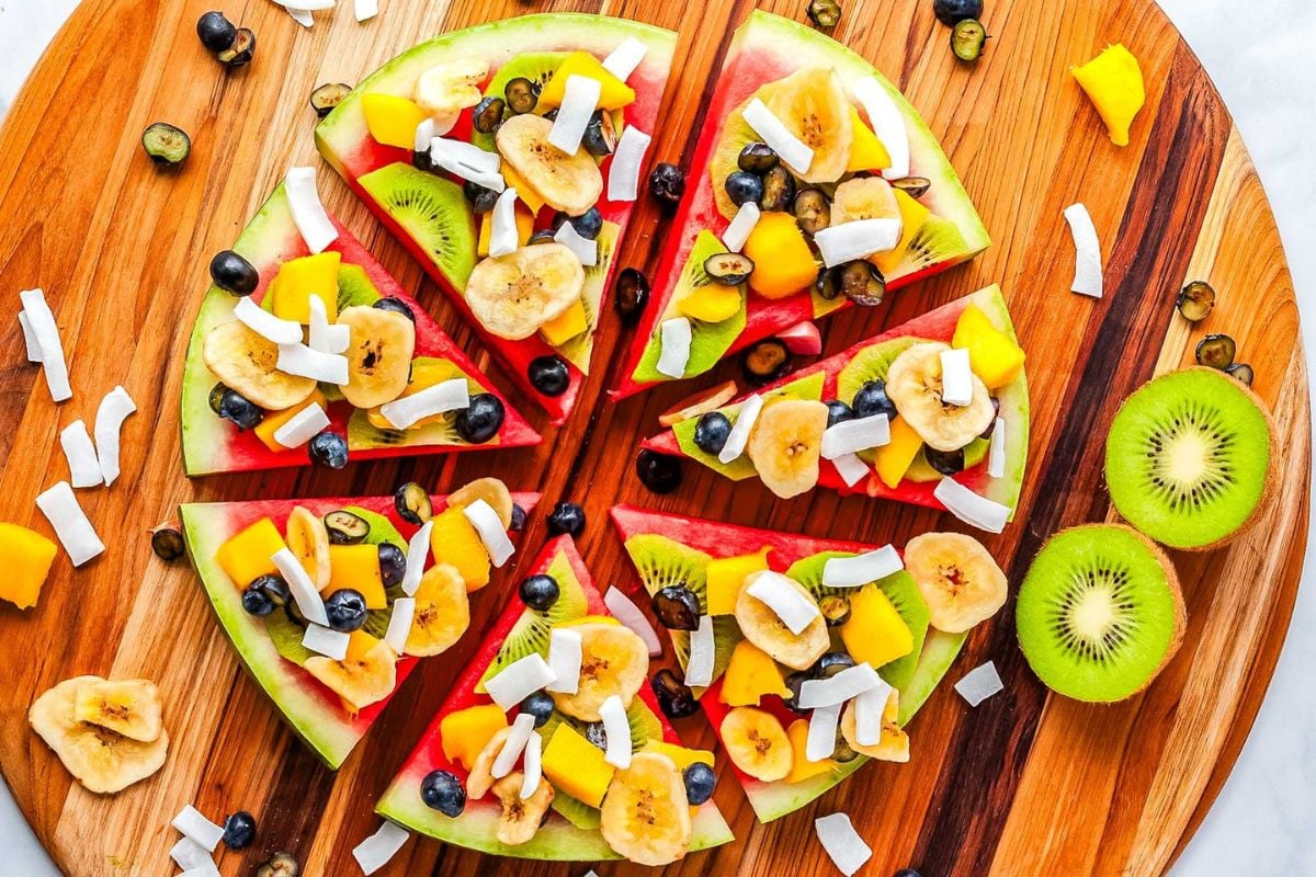Sliced watermelon arranged in a circle, topped with banana chips, kiwi, mango, blueberries, and coconut flakes on a wooden board, with extra fruit scattered around.