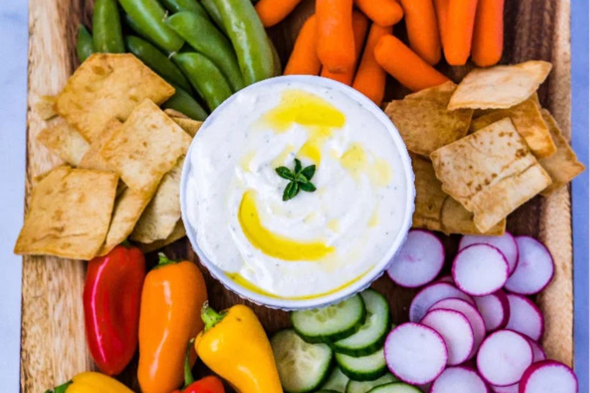 A platter with pita chips, snap peas, carrots, mini bell peppers, cucumber slices, radishes, and a bowl of creamy dip garnished with herbs and olive oil.
