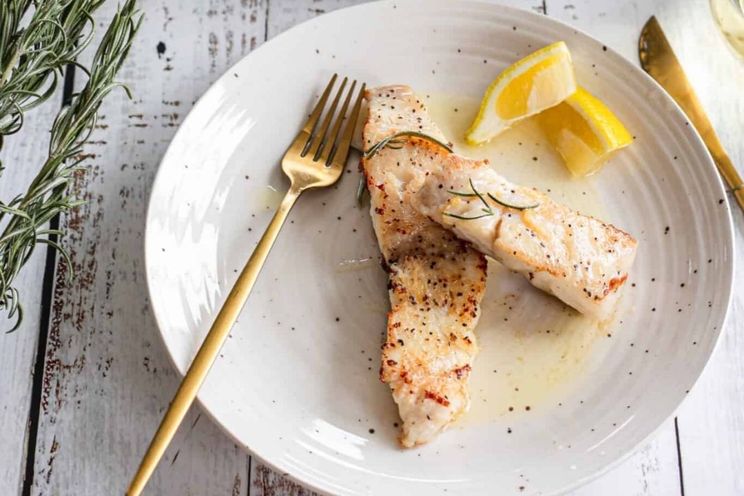 Two pieces of cooked fish garnished with rosemary on a white plate, served with lemon wedges and a gold fork on a rustic white table—perfect for light recipes that make unforgettable seafood dinners on warm summer nights.