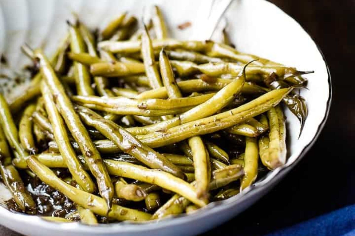A white bowl filled with cooked green beans coated in a dark sauce.