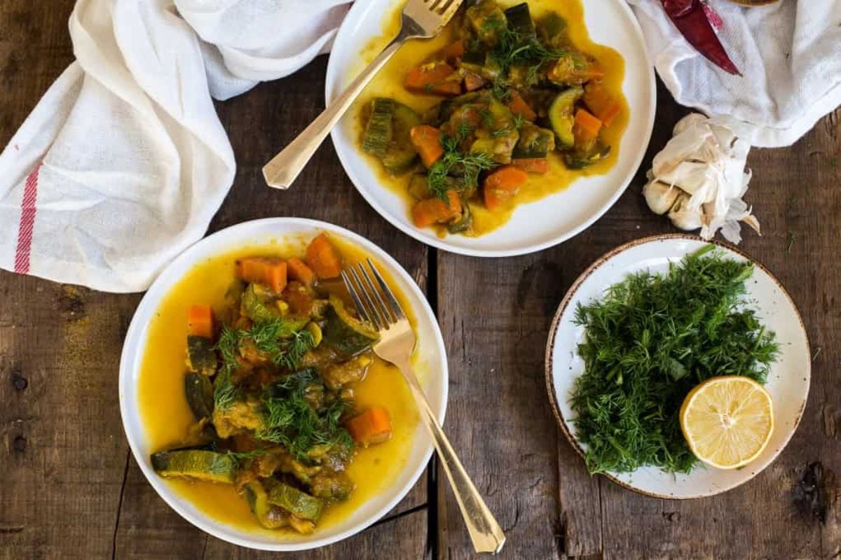Two plates of vegetable stew with fresh herbs on a rustic wooden table, alongside a small plate holding fresh dill and a lemon half.