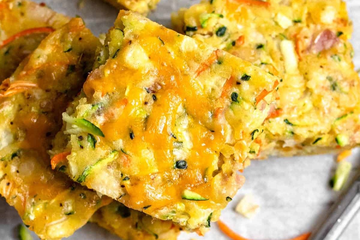 Close-up of cheesy vegetable squares with visible pieces of zucchini, carrots, and melted cheese on top, stacked on a light surface.
