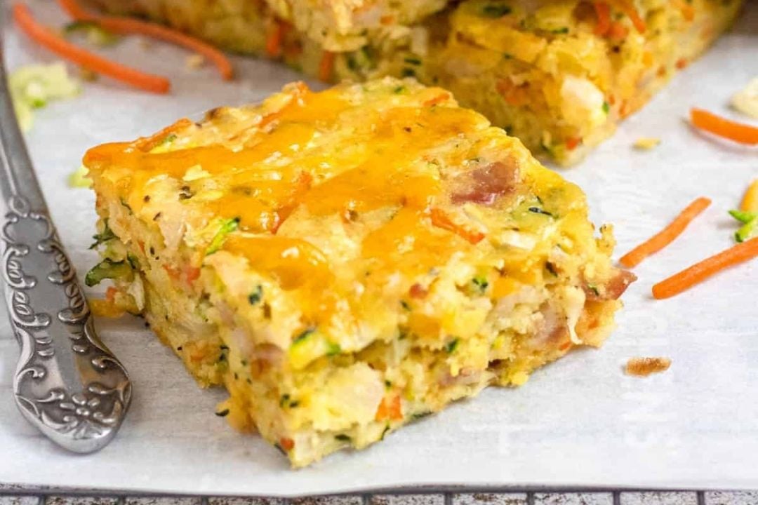 A square piece of cheesy baked casserole with visible bits of vegetables and ham, placed on parchment paper next to a metal fork.