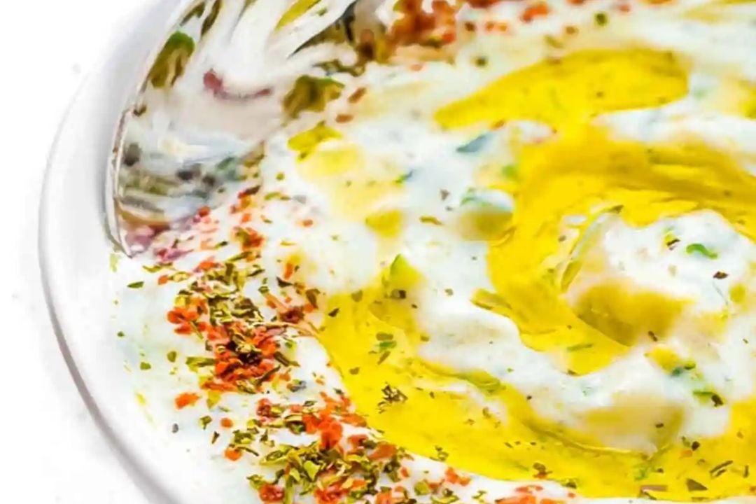 A plate of yogurt topped with olive oil, dried herbs, and red pepper flakes, with a spoon partially visible.