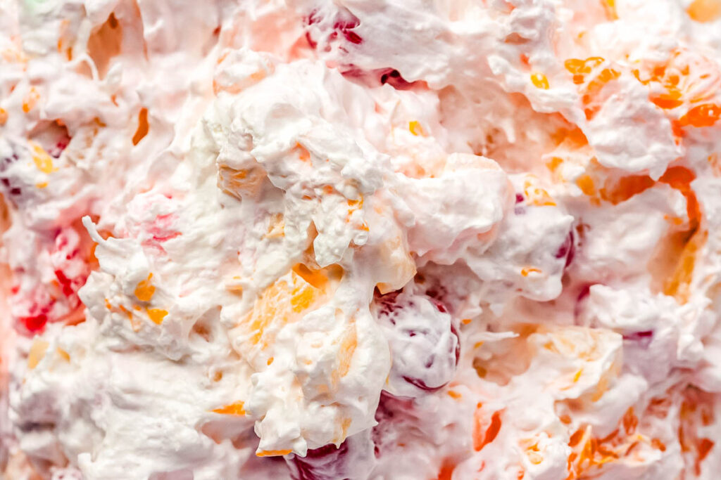 A close-up view of a creamy fruit salad with whipped topping, showing visible pieces of orange, red, and yellow fruit mixed in—perfect for potluck salads or travel friendly salads at your next gathering.