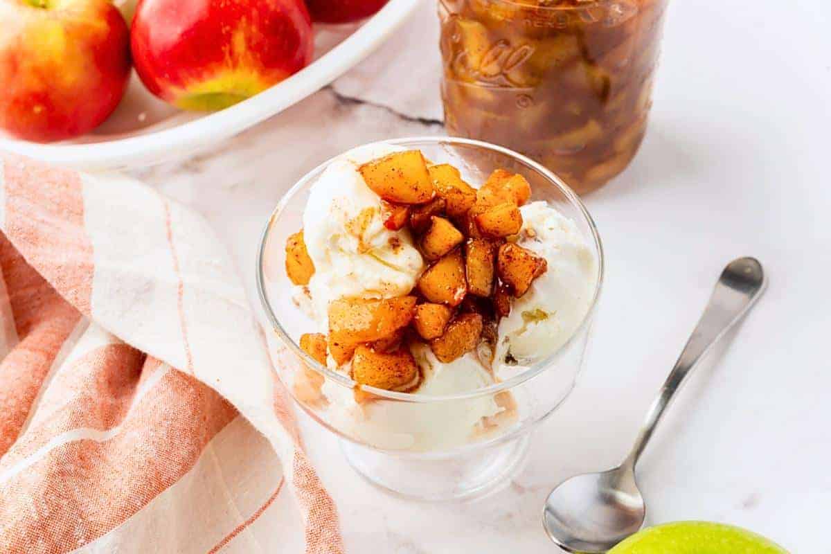 A glass bowl of vanilla ice cream topped with cinnamon-spiced apple chunks, next to a spoon, red apples, and a jar of apple topping on a white surface—a delicious way to celebrate apple season with classic apple recipes.