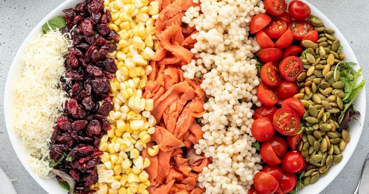 A salad bowl with rows of shredded cheese, dried cranberries, corn, smoked salmon, couscous, cherry tomatoes, and pumpkin seeds on a bed of greens—perfect as one of your make-ahead Labor Day sides for stress-free hosting.