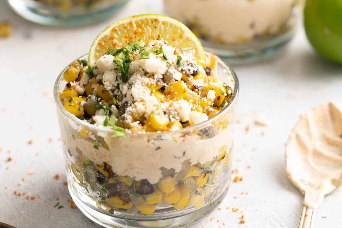 A glass cup filled with layers of corn, creamy sauce, cheese, cilantro, and garnished with a lime slice sits on a light-colored surface—a garden-fresh creation perfect for your collection of summer recipes.