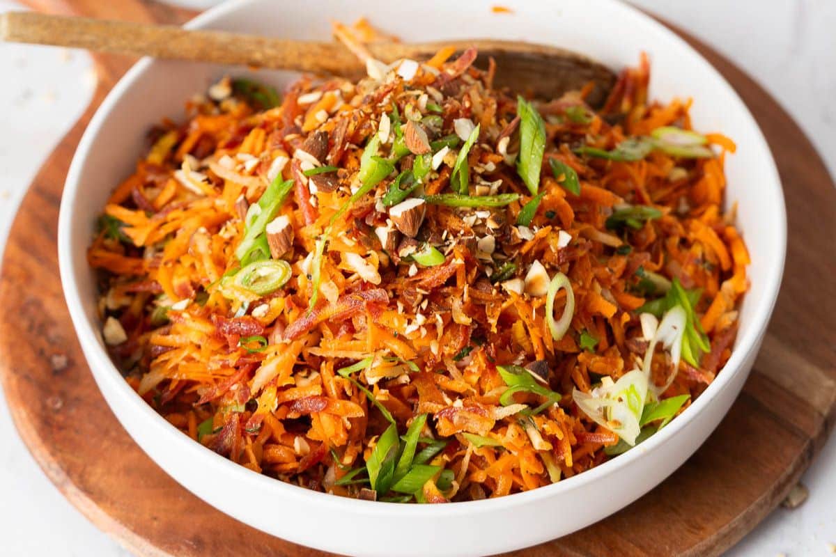 A bowl of shredded carrot salad topped with chopped nuts and sliced green onions, with a wooden spoon resting inside—one of our reader favorites when it comes to popular side dishes.