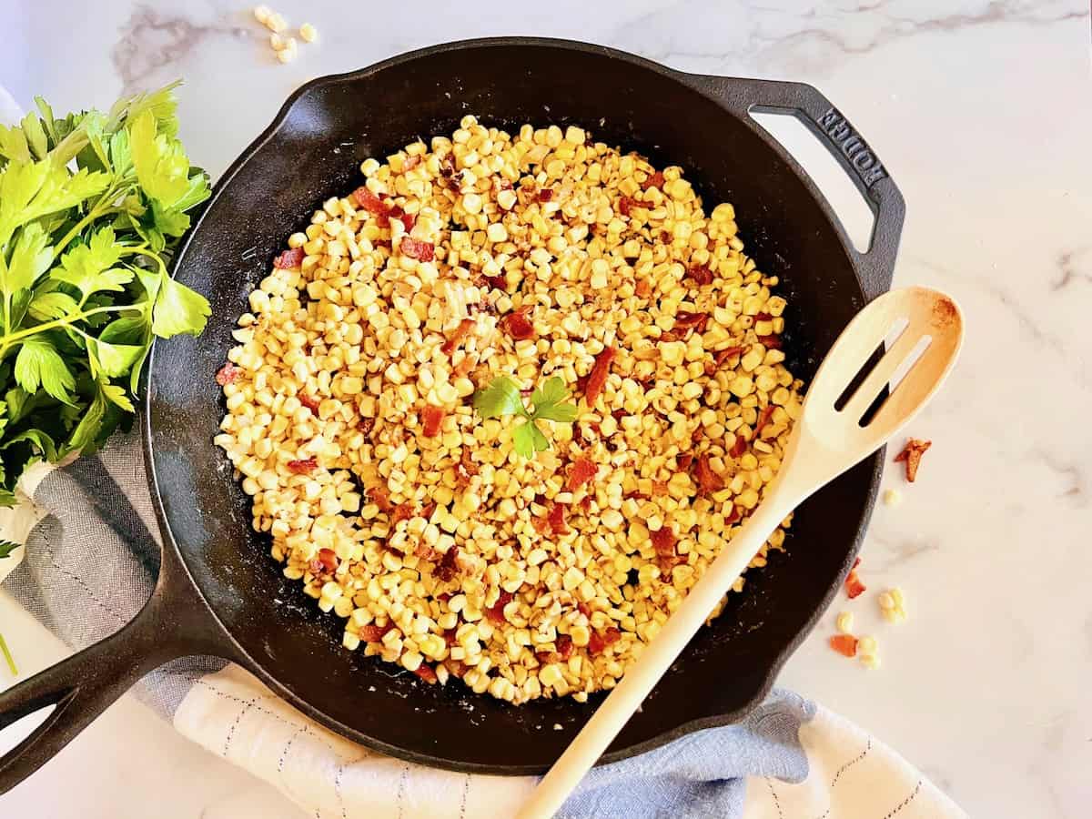 Skillet filled with sautéed corn and bits of bacon, garnished with parsley—an irresistible example of easy vegetable dishes. A wooden spoon rests in the pan, while fresh parsley and a towel are nearby on a marble countertop.
