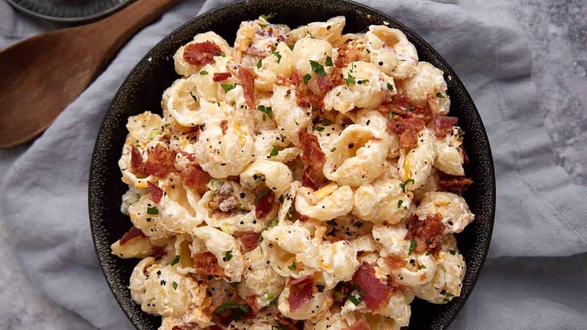 A black bowl filled with creamy pasta shells topped with bacon pieces and chopped herbs, placed on a gray surface with a wooden spoon nearby—this dish is one of those tried-and-true recipes that’s sure to become a favorite at any gathering.