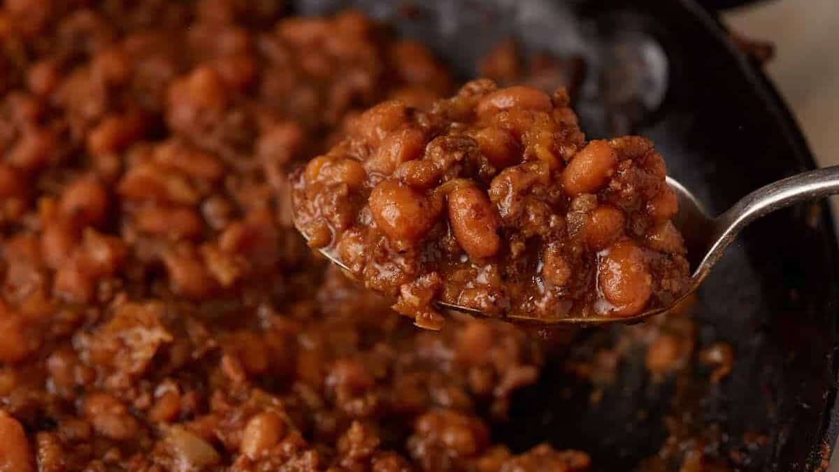A close-up of a spoonful of baked beans and ground meat in a thick, reddish-brown sauce held over a pan of the same mixture—perfect for stress-free hosting and ideal as one of your make-ahead Labor Day sides.