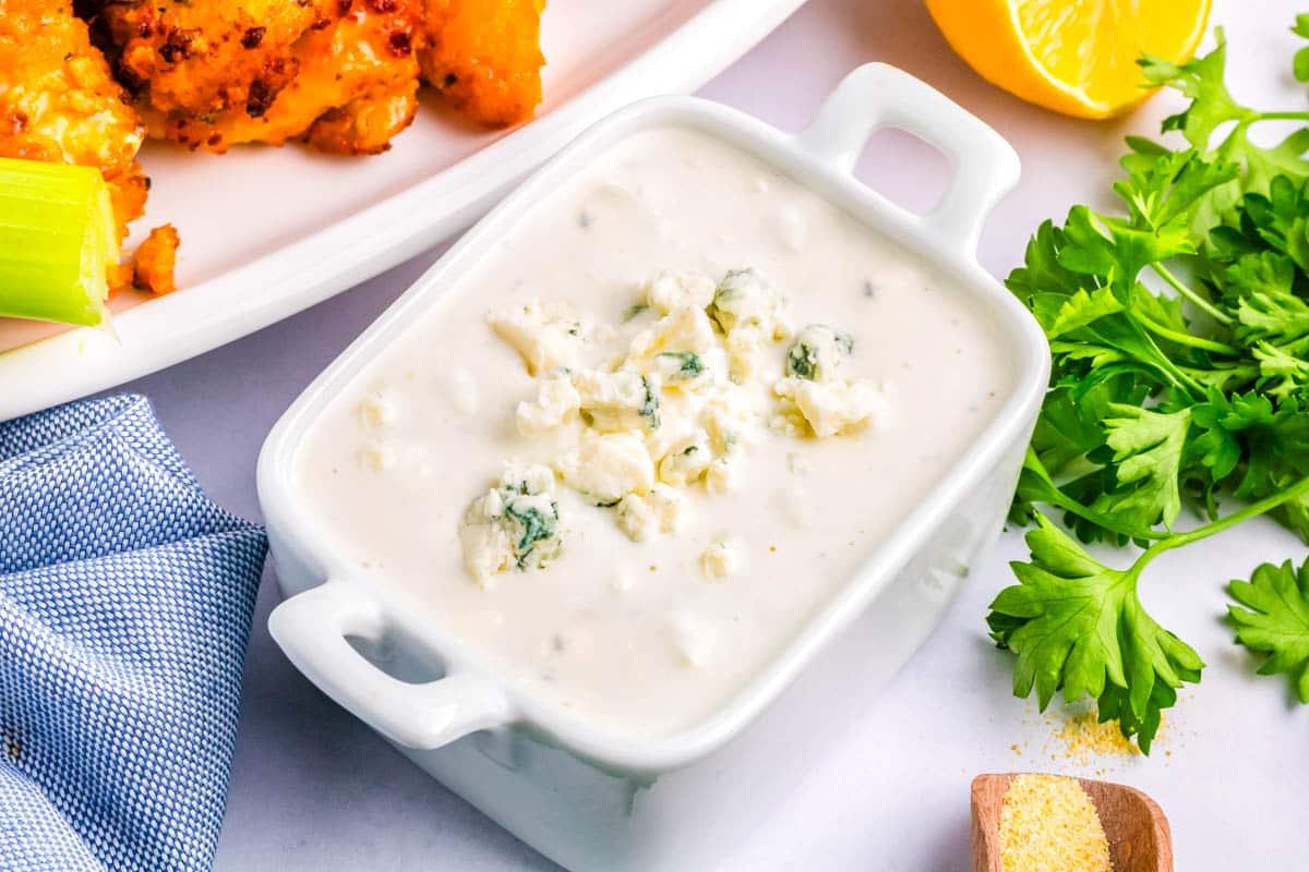 Rectangular dish filled with creamy blue cheese dressing, one of the best dressings for wings or salads, garnished with blue cheese crumbles, surrounded by parsley, lemon, and a plate of buffalo wings.