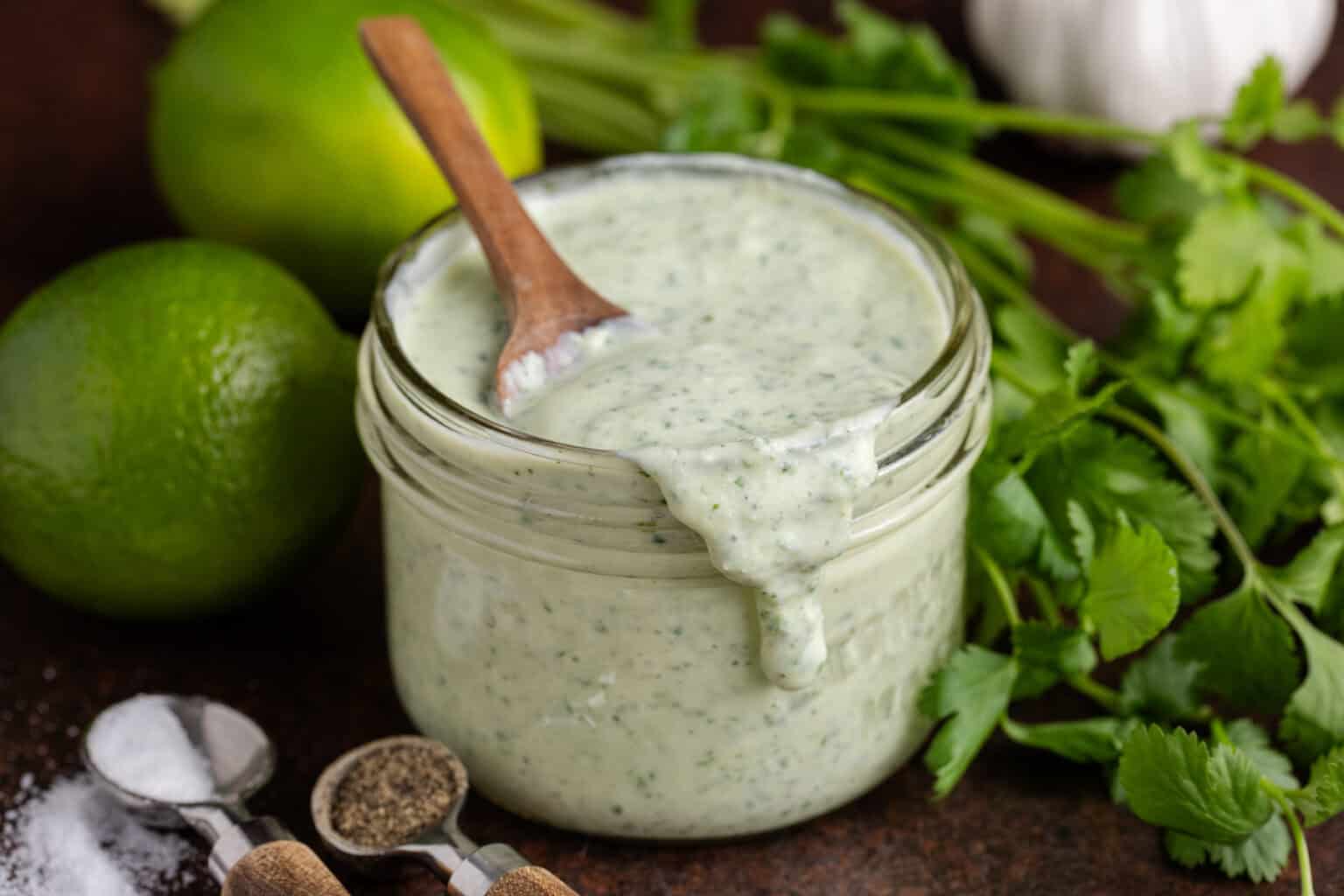 A glass jar filled with creamy green sauce, with a small wooden spoon inside. Fresh cilantro, limes, garlic, salt, and pepper are arranged around the jar—perfect inspiration for homemade dressings or to add to your favorite dressing recipes.