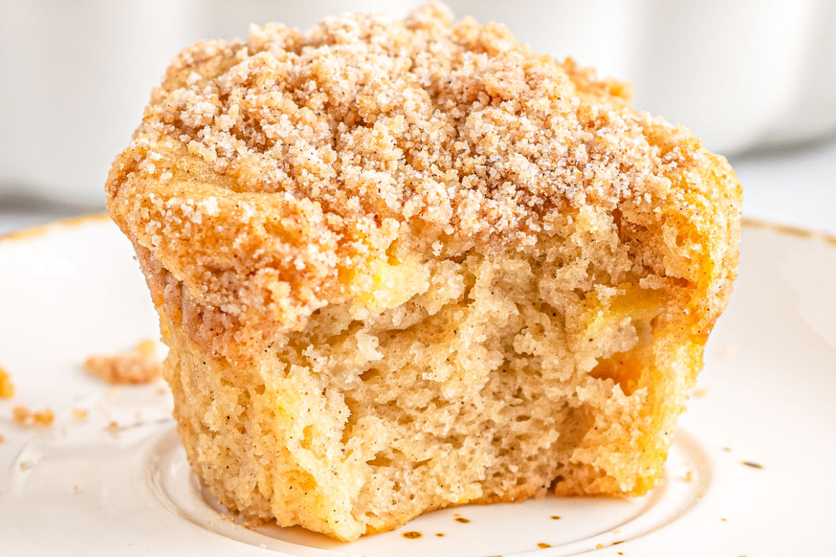 A close-up of a partially eaten crumb-topped muffin on a white plate, showing a light, fluffy interior with visible crumbs around it—an inviting treat perfect for apple season and those who love delicious seasonal recipes.