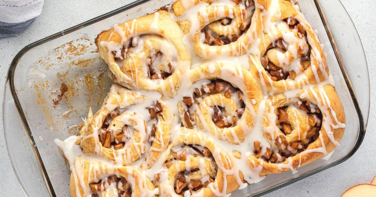 A glass baking dish with eight frosted cinnamon rolls, one missing from the corner, viewed from above on a light countertop—an inviting treat perfect for apple season and those who love cozy seasonal recipes.