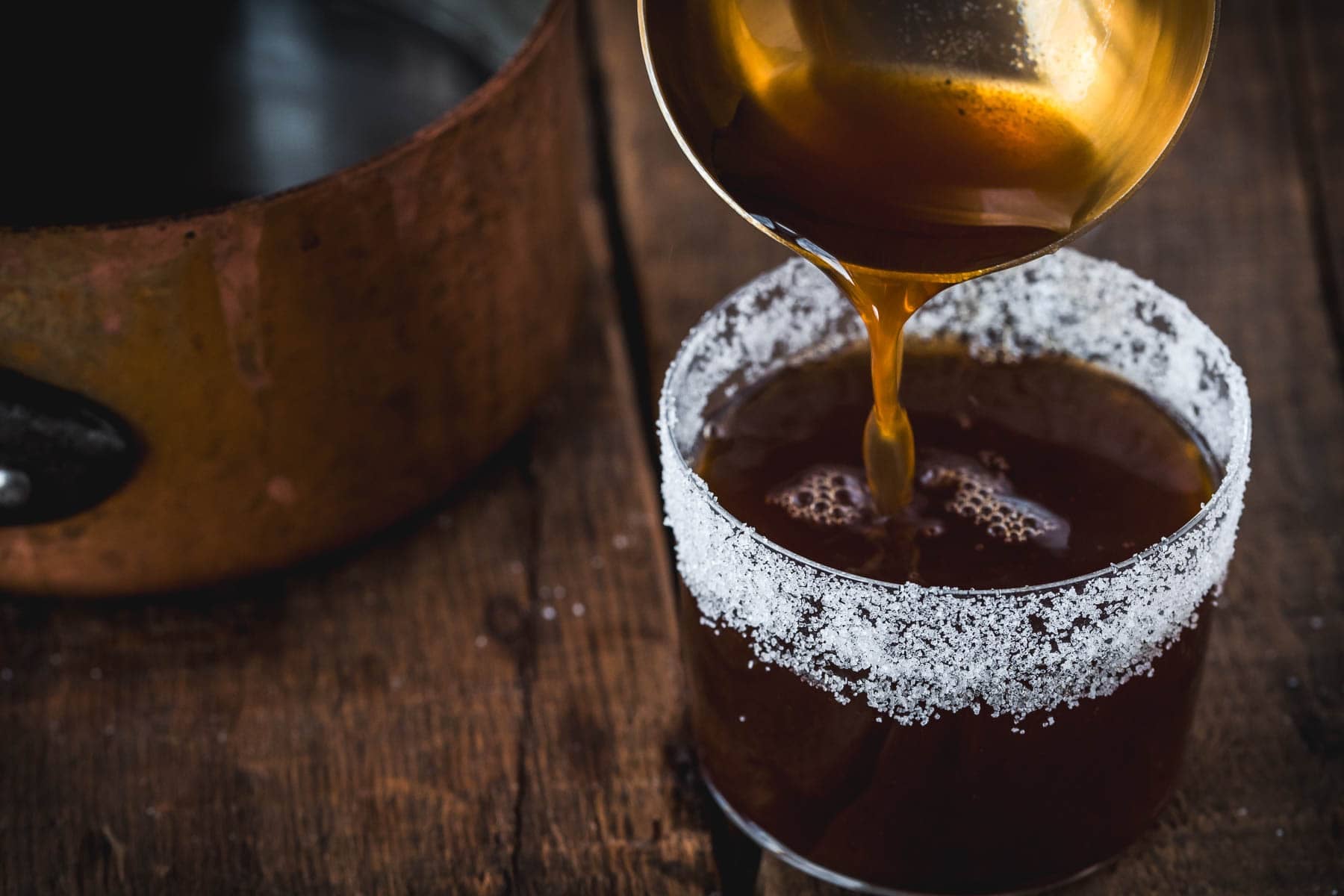 A ladle pours dark liquid into a glass rimmed with sugar on a wooden surface, with a copper pot in the background—an inviting scene perfect for those seeking cozy campfire cocktails.