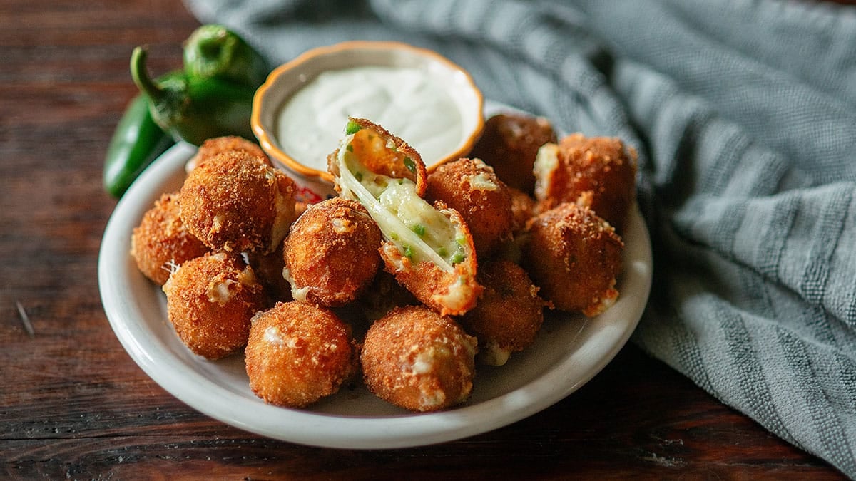A plate of breaded, fried jalapeño poppers with creamy dip and whole jalapeños sits on a wooden table with a gray napkin—perfect for serving as Game Day Appetizers that are sure to spice up your next gathering.