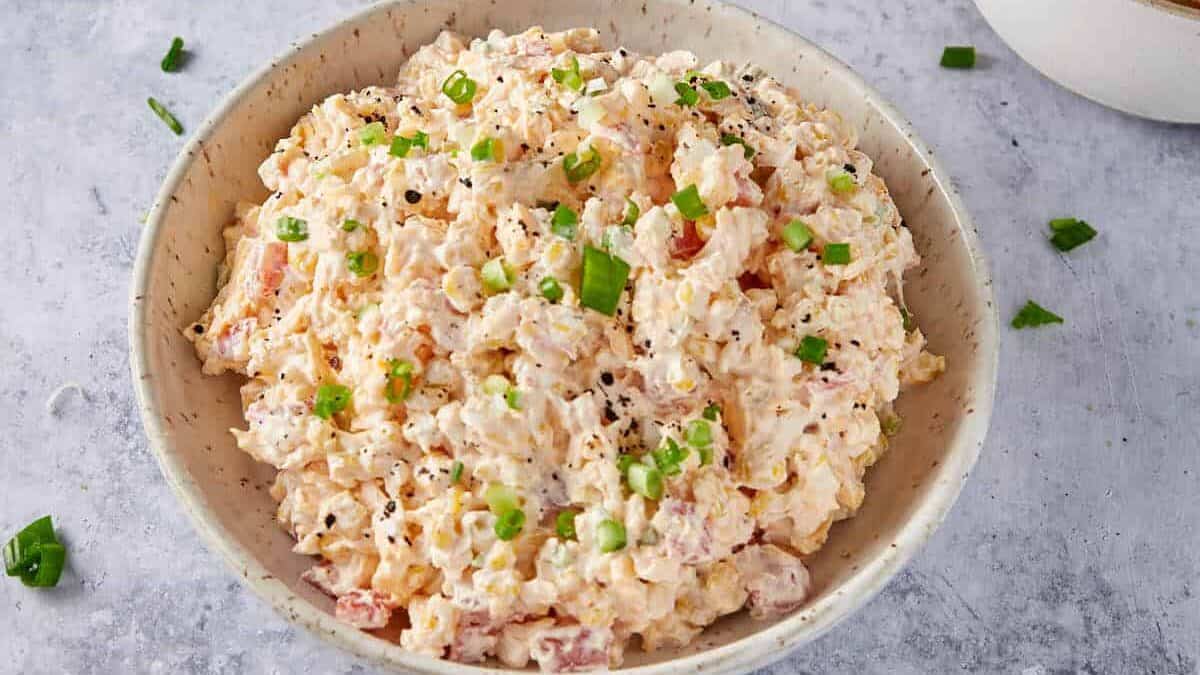 A bowl of creamy pimento cheese dip, perfect for snacking or serving as a crowd-pleasing game day snack, is topped with chopped green onions and black pepper and presented on a light gray surface.