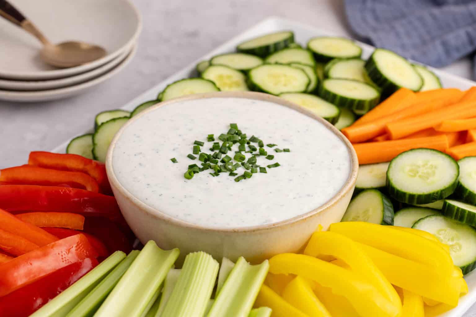 Bowl of ranch dip garnished with chopped chives, surrounded by sliced cucumbers, carrots, red and yellow bell peppers, and celery sticks on a platter—perfect for snacking or serving as a refreshing cold dip at your next game day gathering.