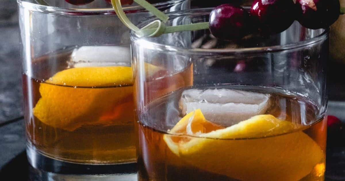 Two glasses of whiskey with large ice cubes, an orange peel garnish, and skewered dark cherries on top—perfect outdoor cocktails to enjoy by the fire pit.