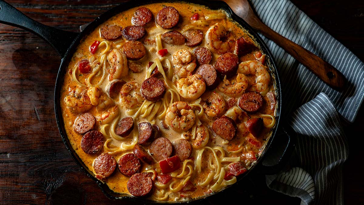 A skillet filled with creamy pasta, shrimp, sliced sausage, and chopped red peppers sits on a wooden surface next to a striped cloth and wooden spoon—perfect inspiration for cozy pasta dinners or new dinner ideas.