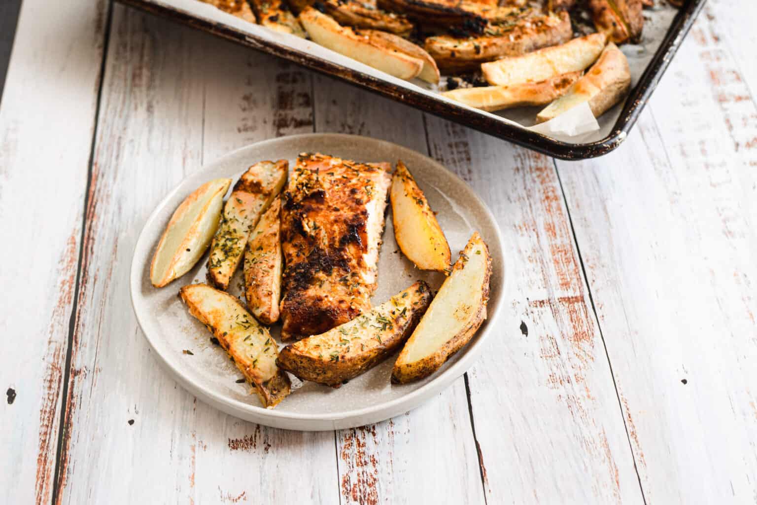 A plate with seasoned baked potato wedges and a piece of spiced roasted chicken, next to a tray with more wedges on a rustic white wooden table—perfect for easy dinners or quick weeknight meals.