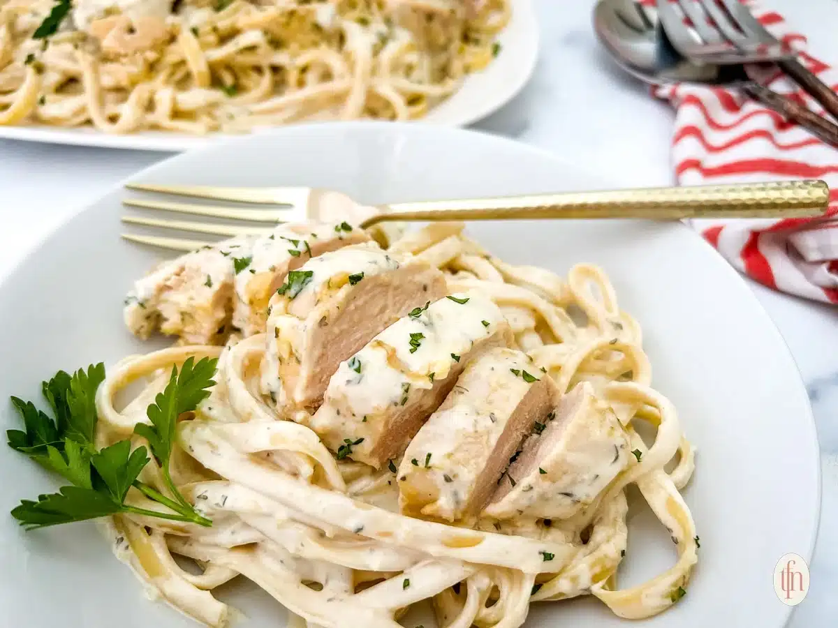 A plate of fettuccine pasta topped with sliced chicken breast in a creamy white sauce, garnished with parsley, with a fork on the side—this dish is perfect for comforting fall recipes and can easily be adapted for your slow cooker.
