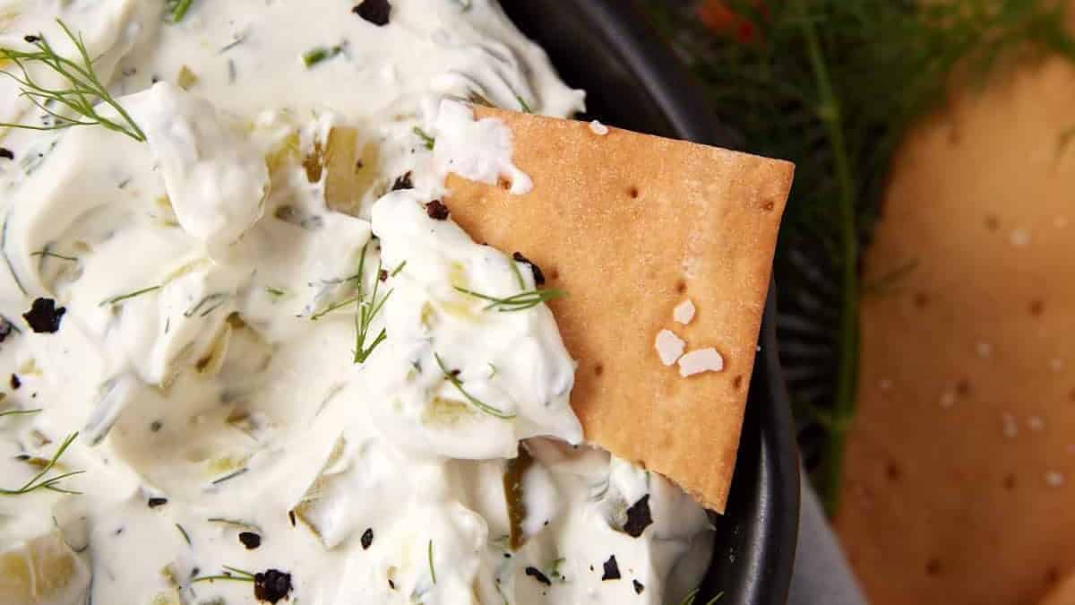 A cracker dipped into a creamy white dip with herbs and black pepper, served in a dark bowl—perfect for snacking or serving as one of your go-to cold dips on game day.
