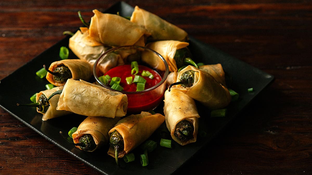 A black plate with stuffed, rolled appetizers—perfect as game day appetizers—garnished with chopped green onions and arranged around a bowl of red dipping sauce.