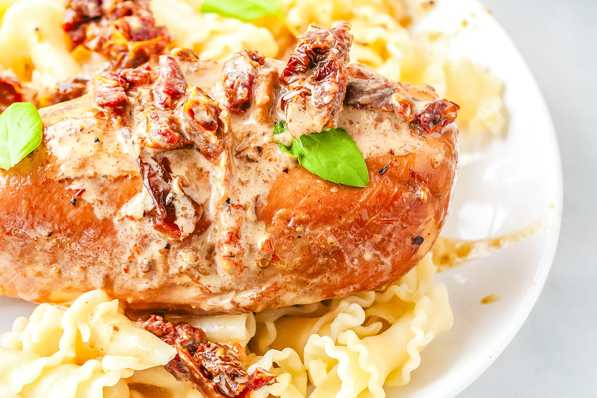 Cooked chicken breast topped with creamy sauce, sun-dried tomatoes, and basil served over ribbon pasta on a white plate—an inviting dish that’s perfect for fall recipes and comforting slow cooker dinners.